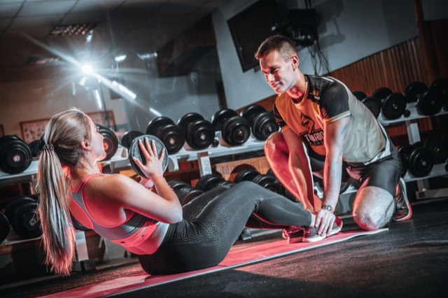 Coach auxiliando aluna em exercício abdominal com anilha